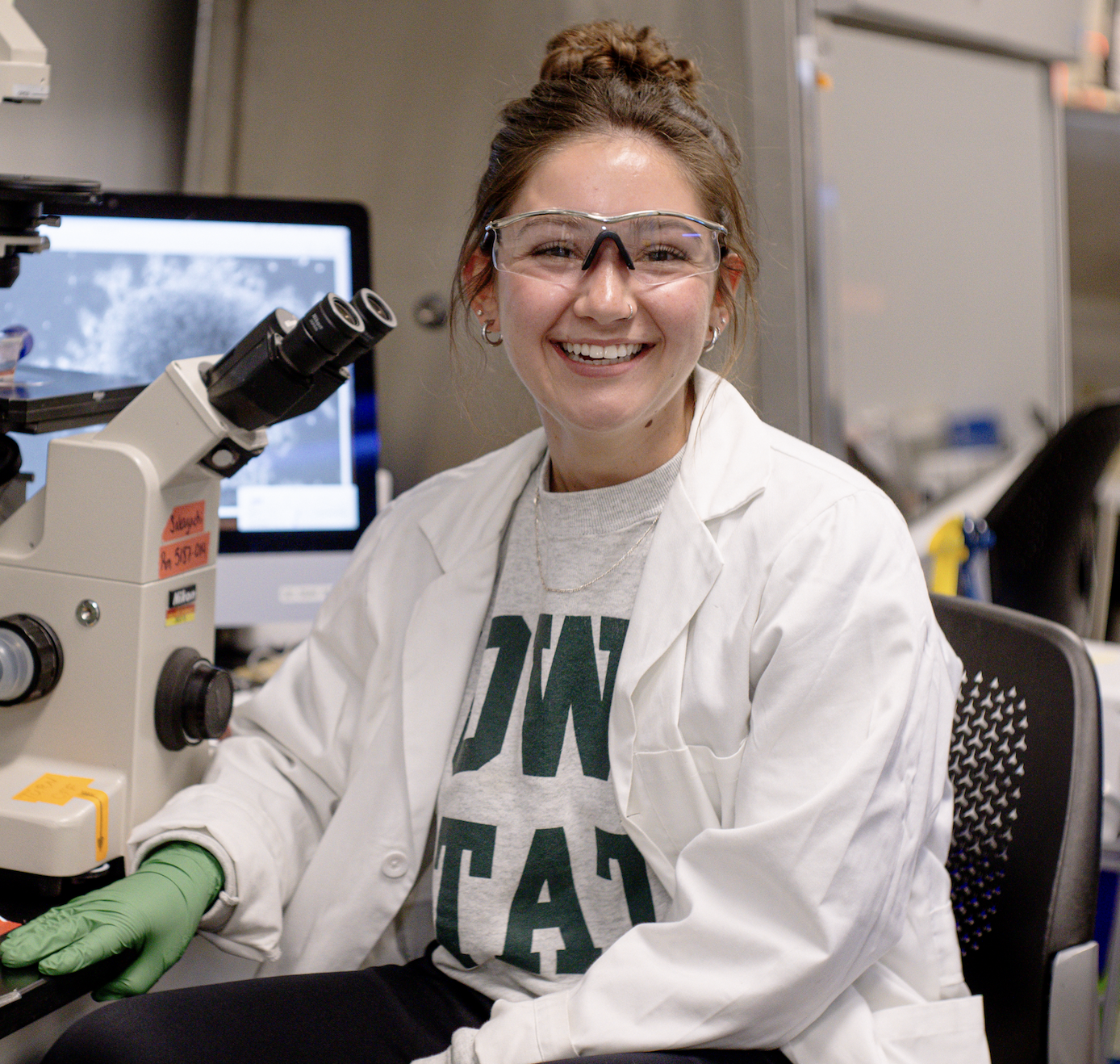 Bridget McGovern, senior in Biology, smiling at the camera wearing protective eye wear and a white lab coat in the Sakaguchi Lab near a microscope.